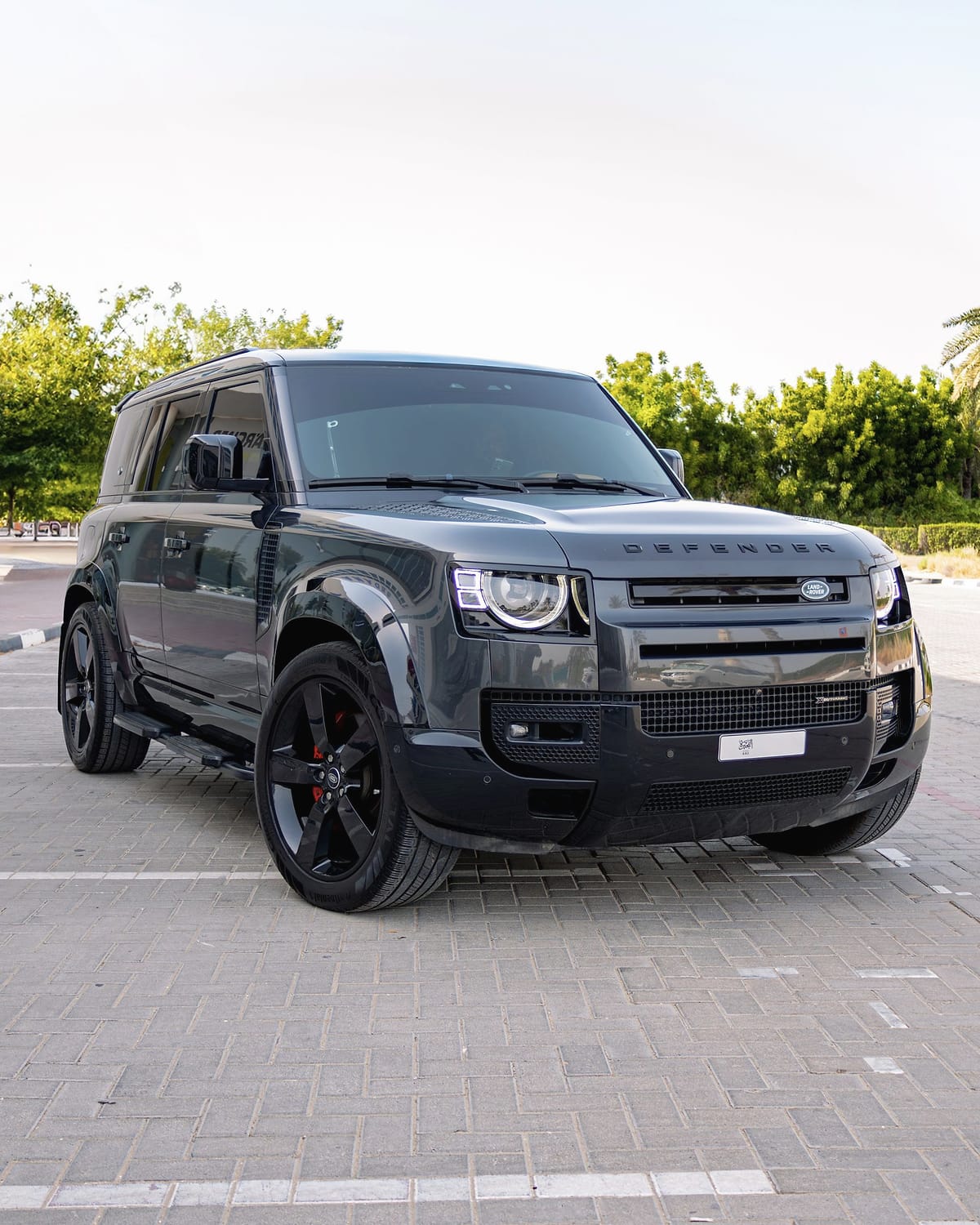 A dark grey Land Rover Defender 110 on pale paving under palms.